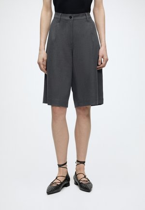 Femme portant un short gris foncé sur mesure longueur genou et des chaussures plates noires à lacets avec des clous argentés sur fond blanc.