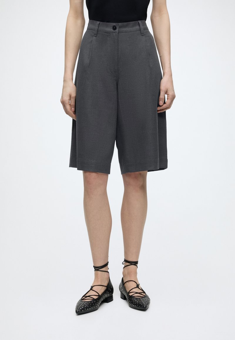 Femme portant un short gris foncé sur mesure longueur genou et des chaussures plates noires à lacets avec des clous argentés sur fond blanc.