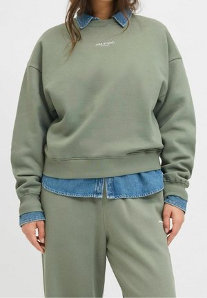 Persoon die een olijfgroene trui en sweatpants draagt over een blauw denim overhemd, staand tegen een effen achtergrond.