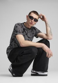 Sort-hvid grafisk T-shirt med gentagende tekstmønster, parret med sorte løse jeans og sort-hvide sneakers. Runde solbriller båret.