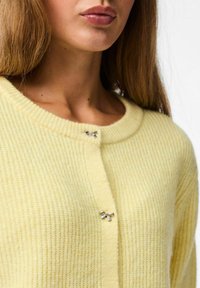 Dzeltens adīts kardigans ar ribotu tekstūru, ar noapaļotu dekoltē un sudraba loku akcentu. Redzami pogu aizdari.