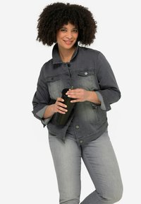 Veste en denim gris avec des boutons et deux poches poitrine, associée à un jeans gris clair. Tient une tasse noire texturée avec une paille.