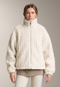 Vit fleecejacka med hög krage, texturerad flätad mönster och dragkedjestängning. Har överdimensionerade ärmar och en kortad fåll.