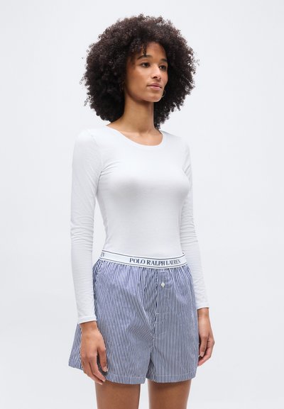 Femme aux cheveux bouclés portant une chemise blanche à manches longues et un short Polo Ralph Lauren à rayures bleues et blanches, debout devant un fond uni.