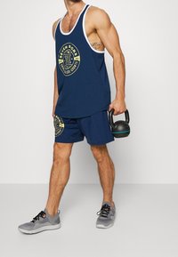 Granatowy podkoszulek z białymi wykończeniami, ozdobiony graficznym logo. W zestawie z granatowymi szortami, trzyma kettlebell w kolorze czarnym i nosi szare buty sportowe.