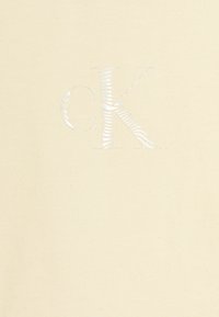 Tessuto di cotone beige con logo "CK" in rilievo di colore bianco lucido, con una texture liscia e un design minimalista.