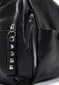 Sac en cuir noir avec une surface texturée, grande fermeture éclair en métal argenté, poche latérale zippée et clous décoratifs en argent sur la sangle.