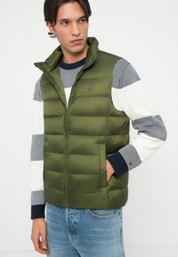 Vest puf olive cu guler înalt, fermoar frontal și două buzunare laterale, purtat peste un pulover tricotat cu dungi și blugi.