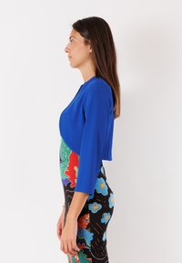 Giacca blu cropped con maniche lunghe indossata sopra un abito floreale multicolore caratterizzato da una silhouette aderente e motivi vivaci su uno sfondo nero.