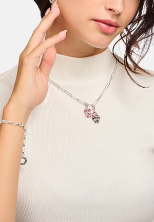 Silberne Kettenhalskette mit einem anhänger im Feiertagsdesign mit roten Akzenten und einem kleinen Charme. Getragen über einem gerippten, cremefarbenen Rollkragenpullover.