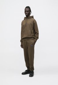 Barna pulóver és melegítő szett puha anyagból, kenguruzsebbel, húzózsinórokkal és kék márkajelzéssel. A kinézetet fekete sportcipő egészíti ki.