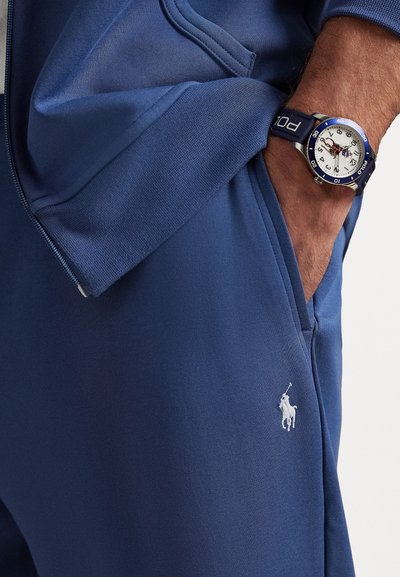 Σακοφτάνα σκούρου μπλε χρώματος και παντελόνι από μαλακό ύφασμα, με λογότυπο κεντημένο σε λευκό χρώμα και ρολόι βασιλικού μπλε με ασημί και λευκό καντράν.