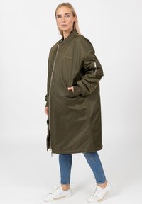 Olijfgroene lange bomberjack met een ritssluiting aan de voorkant, geribbelde manchetten, zijzakken en een gladde, glanzende textuur. Voorzien van een subtiel logodetail.