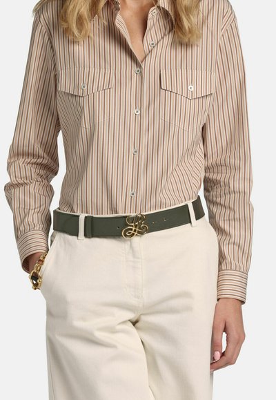 Femme portant une chemise beige à rayures verticales blanches rentrée dans un pantalon blanc cassé avec une ceinture vert foncé à boucle dorée et un bracelet doré.