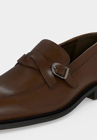 Καφέ δερμάτινο ανδρικό μοκασίνι με ασημένια αγκράφα και μαύρη σόλα, εμφανίζεται από το πλάι σε λευκό φόντο.