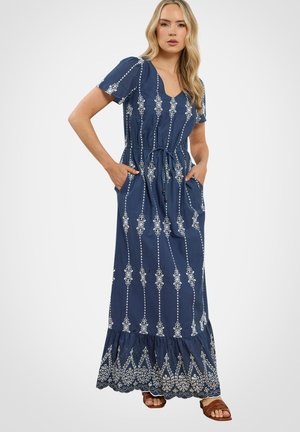 Kobieta stoi, trzymając ręce w kieszeniach, ubrana w granatową sukienkę maxi z białą, pionową haftowaną dekoracją i falbaną u dołu, do tego brązowe sandały.