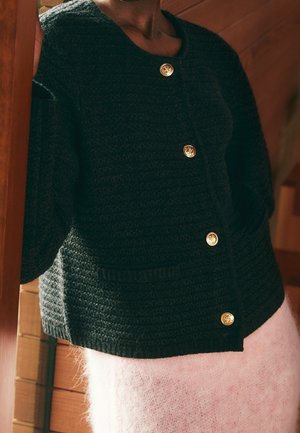 Person trägt eine schwarze Strickjacke mit goldenen Knöpfen und einen hellrosa flauschigen Rock und steht an einem hölzernen Türrahmen.