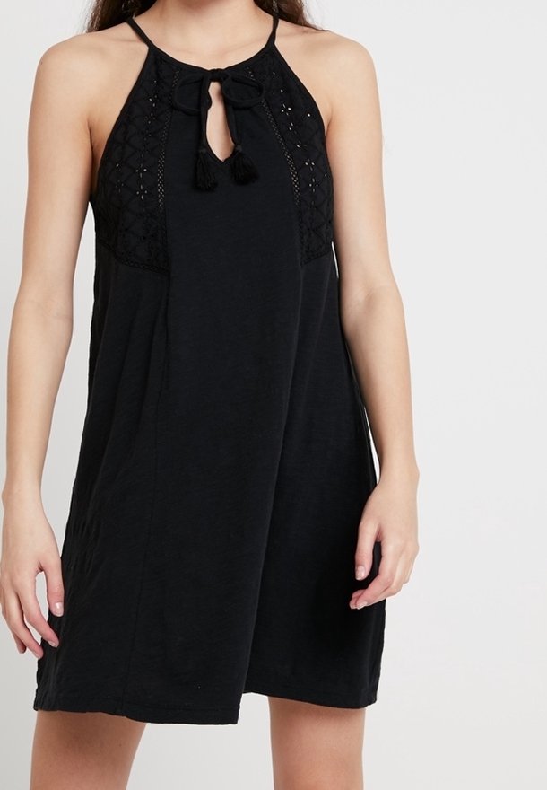 Robe noire sans manches avec des détails en crochet et un décolleté en trou de serrure noué avec des cordons à pompons, portée par une personne debout devant un fond uni.