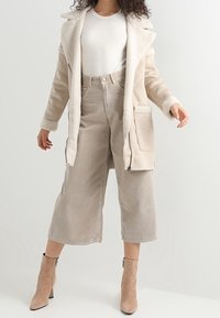 Abrigo de peluche sintético color beige con cuello muesca, combinado con pantalones de pana anchos en beige claro. Botas de tobillo beige completan el conjunto.