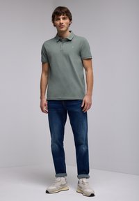 Camisa polo de algodón verde suave, corte clásico, con cuello y mangas cortas. Lleva puesta con jeans azules y zapatillas gris claro.