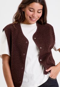 Brown fleece vest with a soft texture, rounded edges, and button closure. Worn over a light-colored shirt, featuring sleeveless design.