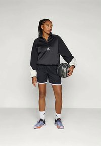 Noor naine mustades spordiriietes hoiab musta Spalding korvpalli, seistes ühe käega lõdvestunult ja vaadates külje poole.