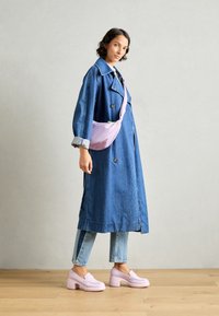 Abrigo de mezclilla azul, largo, con cuello grande y botones. Bolso bandolera rosa claro, vaqueros ajustados y zapatos de plataforma rosa suave. Suelo de madera.