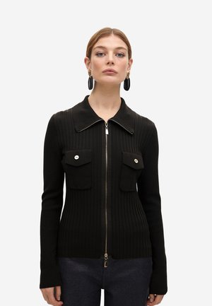 Cárdigan negro de canalé con cremallera en la parte delantera, cuello puntiagudo y dos bolsillos en el pecho. Presenta botones en tono dorado y una textura suave.