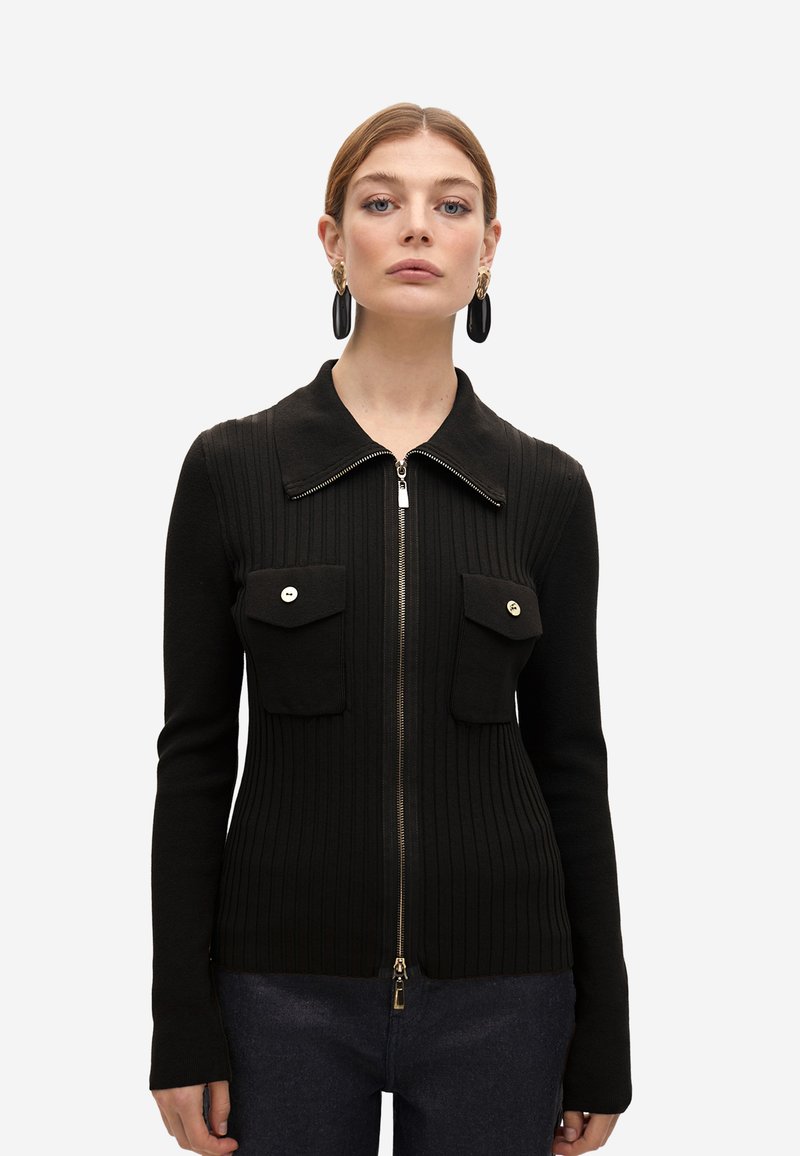 Cárdigan negro de canalé con cremallera en la parte delantera, cuello puntiagudo y dos bolsillos en el pecho. Presenta botones en tono dorado y una textura suave.