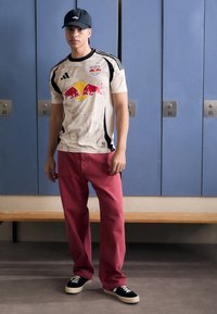 Bílý sportovní dres s černými a červenými akcenty, s logem Red Bull, v kombinaci s volnými červenými džínami a černými teniskami. Pozadí: modré skříňky.