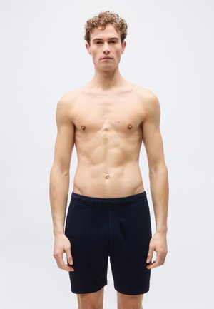 Joven con cabello rizado está de pie sin camiseta, vistiendo pantalones cortos negros texturizados contra un fondo claro y sencillo.