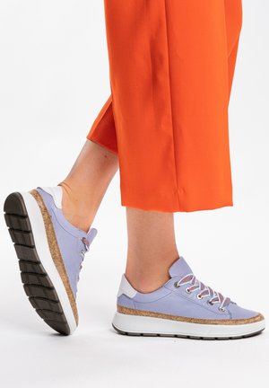 Legs wearing cropped bright orange pants and light purple sneakers with white soles and cork trim, one foot slightly raised.