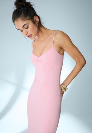 Žena s tmavými vlasmi v drdole, oblečená v ružových bezrukávoch, pozerajúca sa dole, so zlatými náramkami na pravej zápästí na svetlom pozadí.