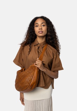 Donna con lunghi capelli ricci che indossa una camicia marrone e una gonna beige, mentre tiene una grande borsa a tracolla in pelle intrecciata marrone.
