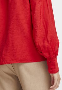 Rote, strukturierte Bluse mit langen, gepufften Ärmeln. Verfügt über knöpfbare Manschetten. Getragen über hellbeigen Hosen. Glatte, geschichtete Stoffoptik.