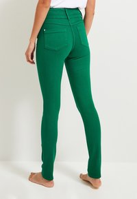 Donna in piedi a piedi nudi, indossa jeans verdi aderenti con tasche posteriori, mostrata dalla vita ai piedi su uno sfondo chiaro e semplice.