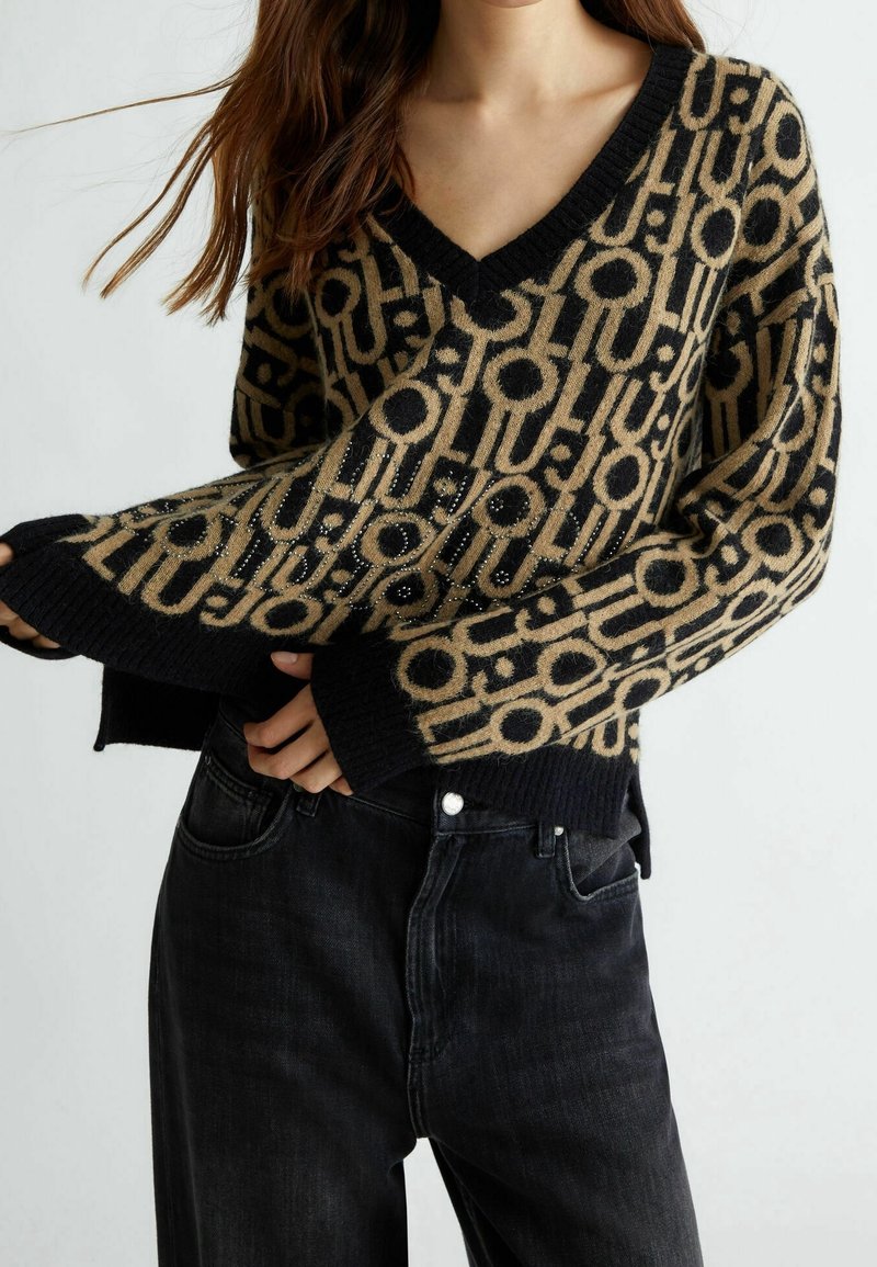Femme portant un pull à col en V à motifs noir et beige avec des poignets côtelés et un jean noir, debout devant un fond uni clair.