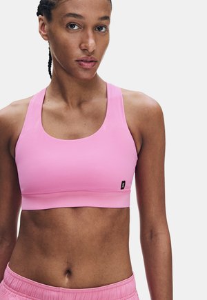 Donna con capelli intrecciati che indossa un reggiseno sportivo rosa e pantaloncini rosa su uno sfondo chiaro uniforme.