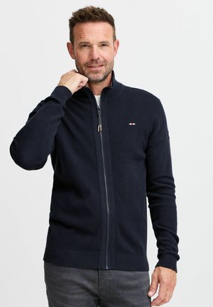 Pulover închis la culoare cu fermoar, textură ribbed și guler înalt. Prezintă un mic logo cu accente roșii, albe și albastre. Design simplu și ajustat.