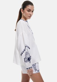 Sæt bestående af en hvid bomuldsblanding top og shorts, prydet med detaljeret mørkeblå broderi og et struktureret mønster. Toppen har en afslappet pasform med lange ærmer.