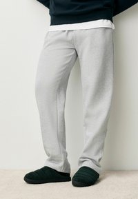 Pantaloni tuta grigi chiari con una texture morbida, caratterizzati da una vestibilità comoda e cuciture laterali. Abbinate a pantofole nere trapuntate su un tappeto beige.