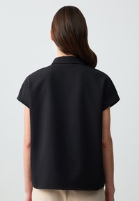 Donna con lunghi capelli castani indossa una maglietta nera a maniche corte e morbida, in piedi con le spalle alla macchina fotografica contro uno sfondo chiaro e semplice.