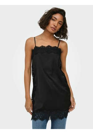 Femme portant un caraco en satin noir avec bordure en dentelle à l'encolure, sur les côtés et à l'ourlet, associé à un jean bleu, sur un fond clair uni.