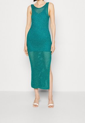 Kobieta w bezrękawowej, turkusowej, szydełkowej sukience midi z bocznym rozcięciem, w zestawieniu z przezroczystymi sandałami na obcasie ozdobionymi perełkami.