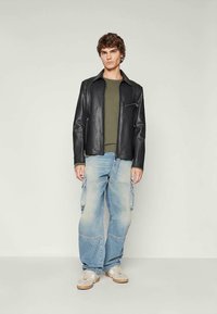 Νεαρός άνδρας που στέκεται φορώντας μαύρο δερμάτινο μπουφάν, ελαιοπράσινη ζακέτα, φωτεινά μπλε cargo jeans και μπεζ αθλητικά παπούτσια σε έναν ουδέτερο φόντο.