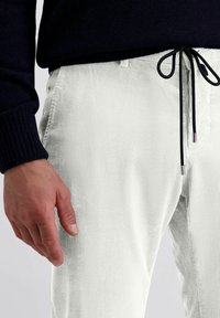 Witte corduroy broek met verticale ribbels, voorzien van zwarte trekkoorden in de taille en een gestructureerd oppervlak, met een gedeeltelijk zichtbare hand aan de zijkant.