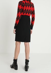Femme portant un pull en argyle rouge et noir, une jupe noire mi-longue avec une fente arrière, et des bottes à talon en cuir verni noires, se tenant contre un fond blanc.