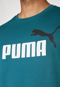 Mėlynas medvilninis marškinėliai su dideliu baltu "PUMA" logotipu ir juodu šuoliuojančio pumos siluetu kairėje pusėje. Lygus paviršius.