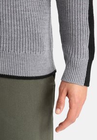 Grauer und schwarzer Strickpullover mit strukturiertem Muster, gerippten Manschetten und kontrastierenden schwarzen Seitenpanelen, kombiniert mit olivgrünen Hosen.