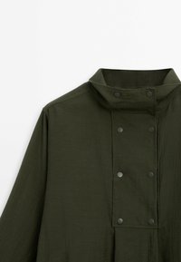 Veste longue à manches longues de couleur vert foncé avec col montant et double rangée de boutons-pression noirs sur le devant.